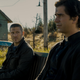 Two men, one dressed as a priest, sit on a bench in a rural setting in Netflix's Midnight Mass.